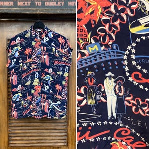 May include: A short-sleeved, button-down shirt with a navy blue background and a colorful Hawaiian print. The print includes palm trees, figures, and the word "Hawaii". The shirt is hanging on a wooden door.