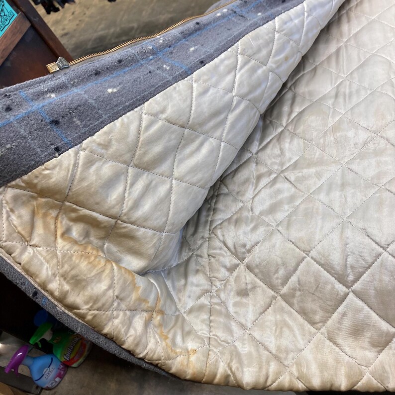 May include: A grey quilted jacket with a zip closure. The lining is white and has a diamond pattern. There are brown stains on the lining.
