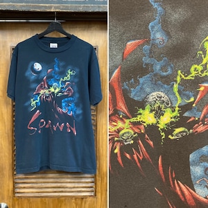 Puede incluir: Una camiseta negra con un estampado gráfico de Spawn, un personaje de cómic, con una capa roja con llamas verdes y azules. El texto "Spawn" está impreso en blanco en la camiseta.