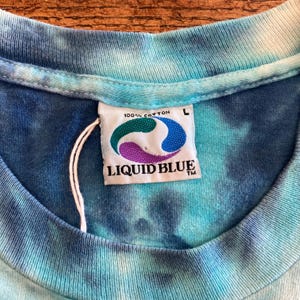 Puede incluir: Primer plano de una camiseta te&ntilde;ida con tintes en tonos azules y turquesas. La etiqueta de la camiseta dice "100% Algod&oacute;n" y presenta un logotipo con formas verdes, azules y moradas, con las palabras "LIQUID BLUE" debajo. La camiseta es talla L.