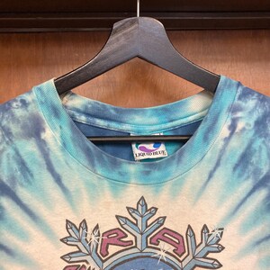 Puede incluir: Una camiseta te&ntilde;ida con un dise&ntilde;o de espiral azul y blanco. La camiseta presenta un dise&ntilde;o gr&aacute;fico con la palabra "RAGA" y formas de copo de nieve. La etiqueta de la marca "LIQUID BLUE" es visible en el cuello.