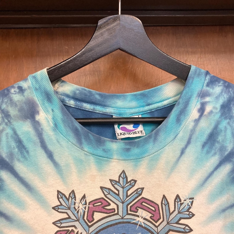 Puede incluir: Una camiseta te&ntilde;ida con un dise&ntilde;o de espiral azul y blanco. La camiseta presenta un dise&ntilde;o gr&aacute;fico con la palabra "RAGA" y formas de copo de nieve. La etiqueta de la marca "LIQUID BLUE" es visible en el cuello.