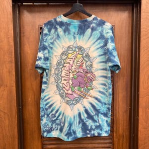 Puede incluir: Camiseta te&ntilde;ida con tinte anudado en azul y blanco con un dise&ntilde;o en espiral. La camiseta presenta un gr&aacute;fico colorido de un personaje de dibujos animados con esqu&iacute;s, rodeado de un dise&ntilde;o floral. Cuelga de una percha negra.