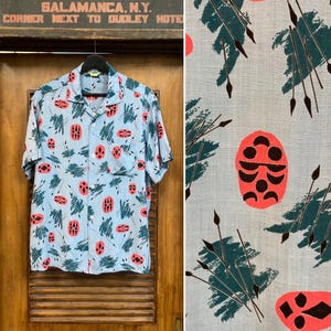 Op de afbeelding: Lichtblauw overhemd met korte mouwen en een uniek patroon. Het overhemd heeft een herhalend ontwerp van koraalkleurige ovale vormen en teal-achtige penseelstrekelementen. Het overhemd hangt aan een zwarte hanger tegen een houten deur.