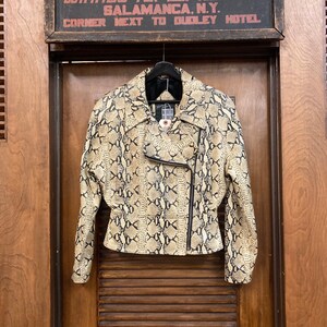 Puede incluir: Una chaqueta de cuero beige y negro con estampado de piel de serpiente y cierre de cremallera. La chaqueta tiene un cuello negro y una cremallera negra en la parte delantera. La chaqueta est&aacute; colgada de una percha.