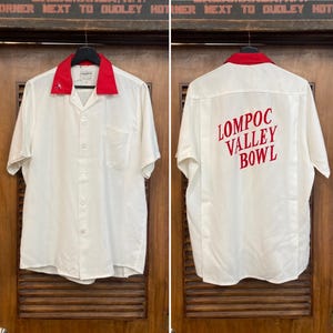 以下が含まれることがあります： 赤い襟と背中に「Lompoc Valley Bowl」の文字が入った白い半袖シャツ。 シャツにはフロントポケットと襟に小さな刺繍デザインがあります。 シャツは木のドアに吊るされています。