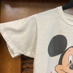 Peut inclure: T-shirt blanc avec un motif de Mickey Mouse. Mickey Mouse porte un short rouge et a des oreilles noires et un nez noir.