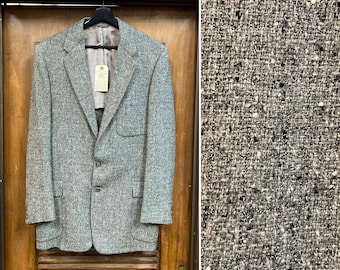 May include: A grey tweed blazer with a three-button fastening, two front pockets, and a notched lapel. The jacket is suspended on a black hanger against a wooden door. A tag is attached to the inside.