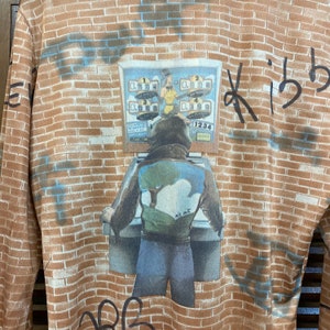 May include: A brown brick wall with a person wearing a brown jacket and blue jeans standing in front of a vintage arcade game. The game has a red, white, and blue design and the text "1234" on the front. The wall has the text "Kiba" written on it.