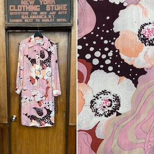 Puede incluir: Un vestido largo de manga larga de color rosa y marrón con estampado floral. El vestido tiene cuello y cierre de botones. La tela tiene un patrón de remolino con flores rosas, marrones, blancas y naranjas.