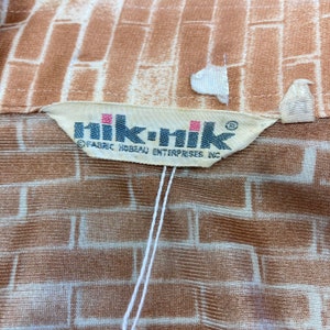May include: A white tag with the text "nik-nik" in black lettering and the text "FABRIC HOBB&Eacute;AU ENTERPRISES INC." in smaller black lettering. The tag is attached to a brown and white fabric with a brick pattern.