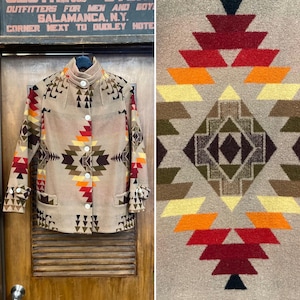 Könnte beinhalten: Beige Jacke mit geometrischem Muster in Rot-, Orange-, Gelb- und Brauntönen. Die Jacke hat einen hohen Kragen, weiße Knöpfe und zwei Vordertaschen. Das Muster wiederholt sich auf den Ärmeln und dem Rücken. Die Jacke hängt an einer Holztür.