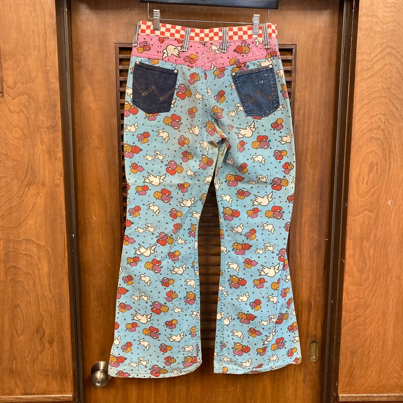 May include: A pair of blue bell bottom jeans with a white dove and red heart pattern. The jeans have a pink and red checkered waistband and denim back pockets.