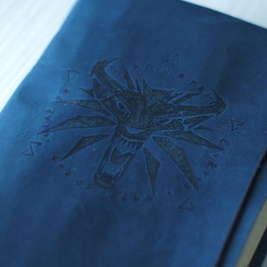 May include: A blue leather journal with a black embossed wolf head emblem. The notebook has a rectangular shape with rounded corners and a visible elastic closure. The pages are cream-colored, and the corners are reinforced with metal.