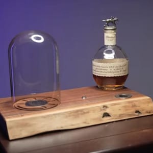 Op de afbeelding: Een houten serveerplank met natuurlijke randen, voorzien van een doorzichtige glazen stolp aan de linkerkant en een glazen fles met een metalen stop in de vorm van een paard aan de rechterkant. Het hout heeft een natuurlijke nerf en knoesten.