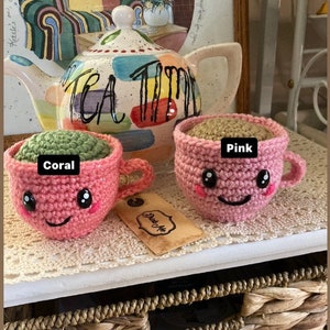 May include: Two crocheted teacups with smiling faces. One cup is coral pink with green tea inside and the other is pink with brown tea inside. The cups are sitting on a wooden surface with a teapot in the background.