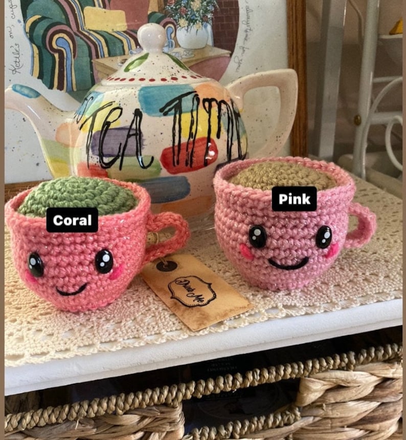 May include: Two crocheted teacups with smiling faces. One cup is coral pink with green tea inside and the other is pink with brown tea inside. The cups are sitting on a wooden surface with a teapot in the background.