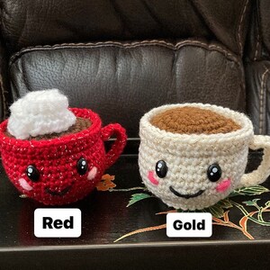 May include: Two crocheted coffee mugs with smiling faces. One mug is red with a white whipped cream topping and the other is gold with brown coffee inside. The mugs are labeled "Red" and "Gold".