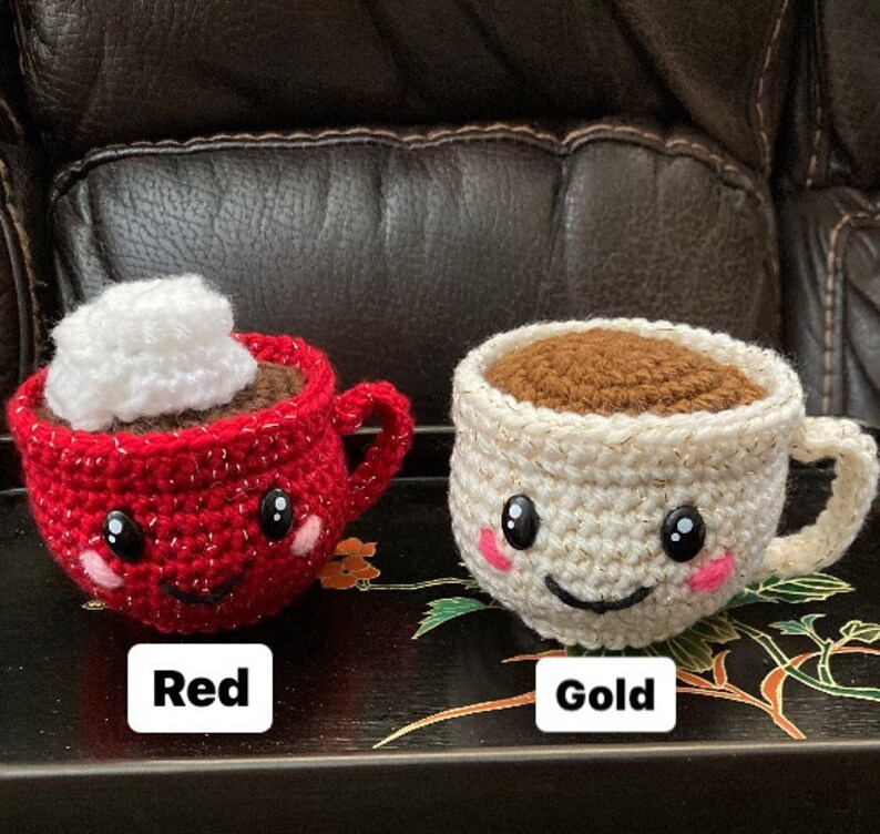 May include: Two crocheted coffee mugs with smiling faces. One mug is red with a white whipped cream topping and the other is gold with brown coffee inside. The mugs are labeled "Red" and "Gold".