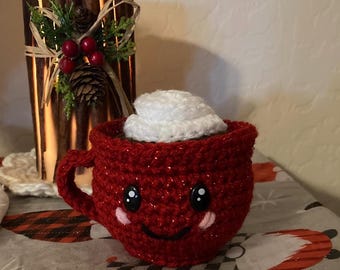 May include: A handmade, red crocheted mug with a smiling face and white whipped cream topping. The mug has a handle and is decorated with black eyes and pink cheeks. A decorative candle holder is in the background.