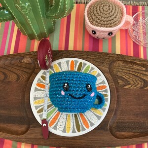May include: A blue crocheted cup with a smiling face and pink cheeks sits on a white and gold patterned plate. A measuring tape and a pink crocheted cup are also in the image.