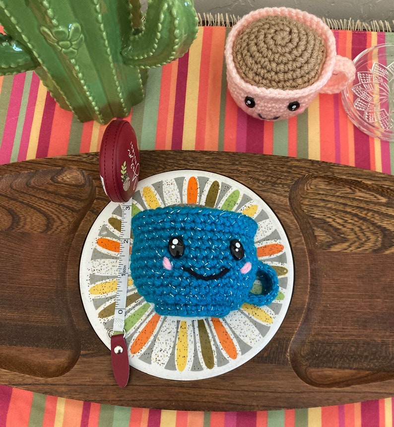 May include: A blue crocheted cup with a smiling face and pink cheeks sits on a white and gold patterned plate. A measuring tape and a pink crocheted cup are also in the image.