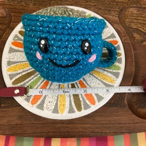 May include: A teal blue crocheted teacup with a smiling face, sparkly accents, and pink cheeks. The teacup is filled with brown yarn and is sitting on a white and gray saucer with a floral design.
