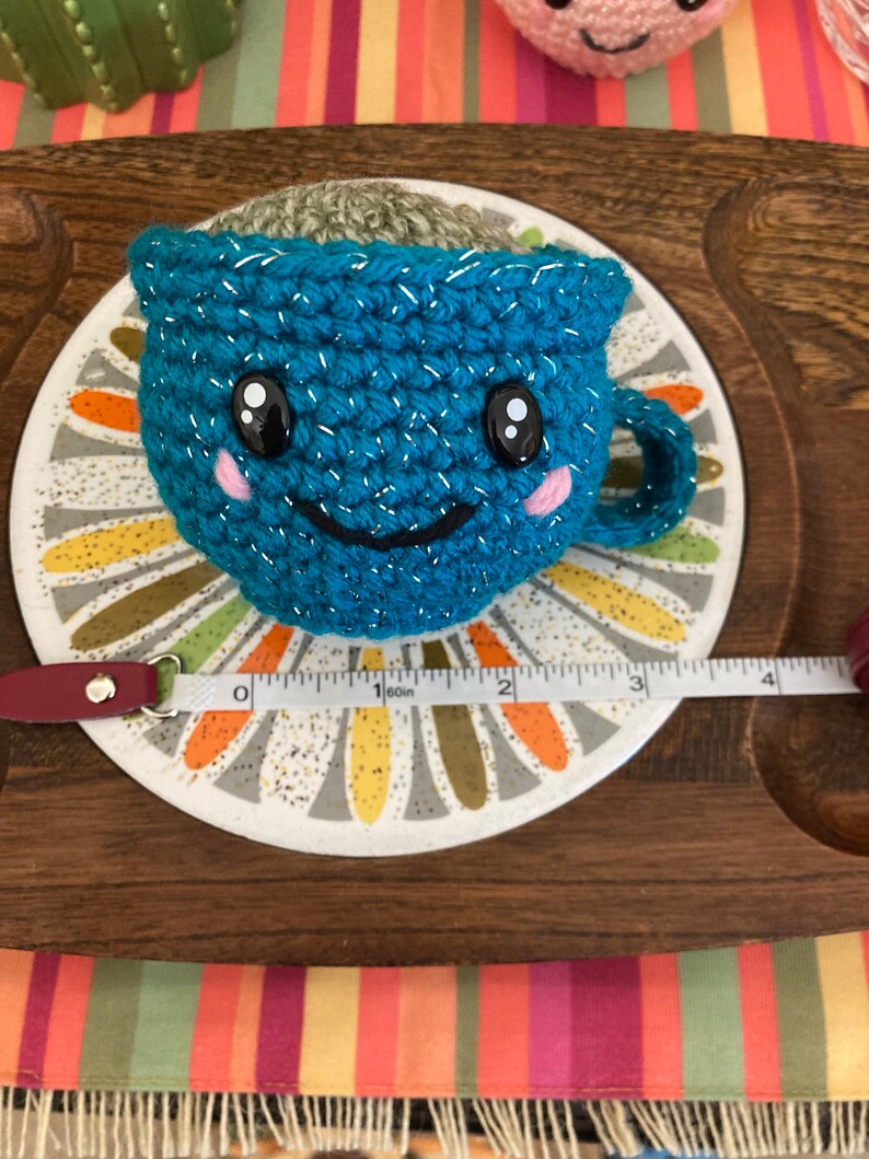 May include: A teal blue crocheted teacup with a smiling face, sparkly accents, and pink cheeks. The teacup is filled with brown yarn and is sitting on a white and gray saucer with a floral design.
