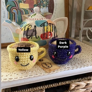 May include: Two crocheted teacups, one yellow and one dark purple, with smiling faces. The cups are filled with brown yarn and have the words "Yellow" and "Dark Purple" written on them. The cups are sitting on a wooden surface with a tea pot in the background.