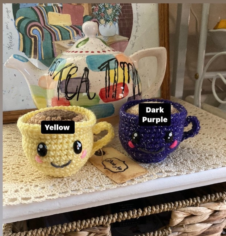 May include: Two crocheted teacups, one yellow and one dark purple, with smiling faces. The cups are filled with brown yarn and have the words "Yellow" and "Dark Purple" written on them. The cups are sitting on a wooden surface with a tea pot in the background.