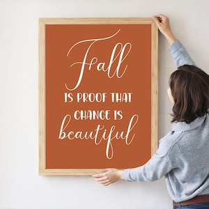 Può includere: Una stampa artistica incorniciata con uno sfondo arancione bruciato e le parole "Fall is proof that change is beautiful" in elegante scrittura bianca. La cornice è color legno chiaro.