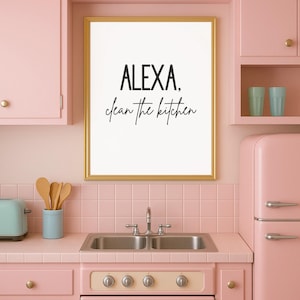 May include: A framed print with the text "ALEXA, clean the kitchen" in a gold frame. The print hangs in a pink kitchen with a retro refrigerator, cabinets, and a double sink.