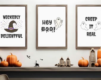 May include: Three framed Halloween-themed prints with text and illustrations. The prints read "Wickedly Delightful", "Hey Boo!", and "Creep It Real". Orange pumpkins and decorative items are arranged on a shelf below the prints.