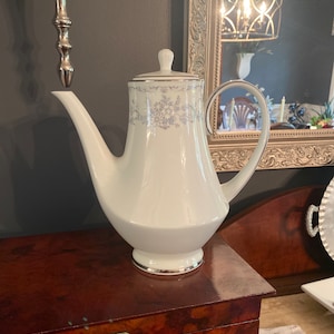 May include: A white porcelain teapot with a silver trim and a floral design in blue and silver. The teapot has a spout and a handle. It is sitting on a wooden surface.