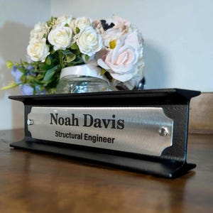 May include: Black desk nameplate with a silver nameplate that reads "Noah Davis, Structural Engineer." The nameplate sits on a wooden desk with a bouquet of flowers in the background.
