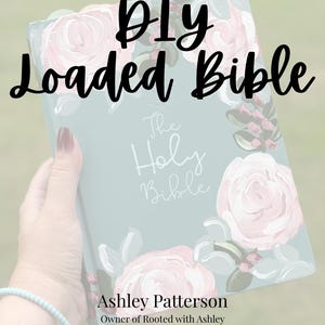 May include: A light blue Bible with a painted floral design featuring pink roses and green leaves. The cover displays the text "The Holy Bible" in white script, with "DIY Loaded Bible" and "Ashley Patterson Owner of Rooted with Ashley" printed.
