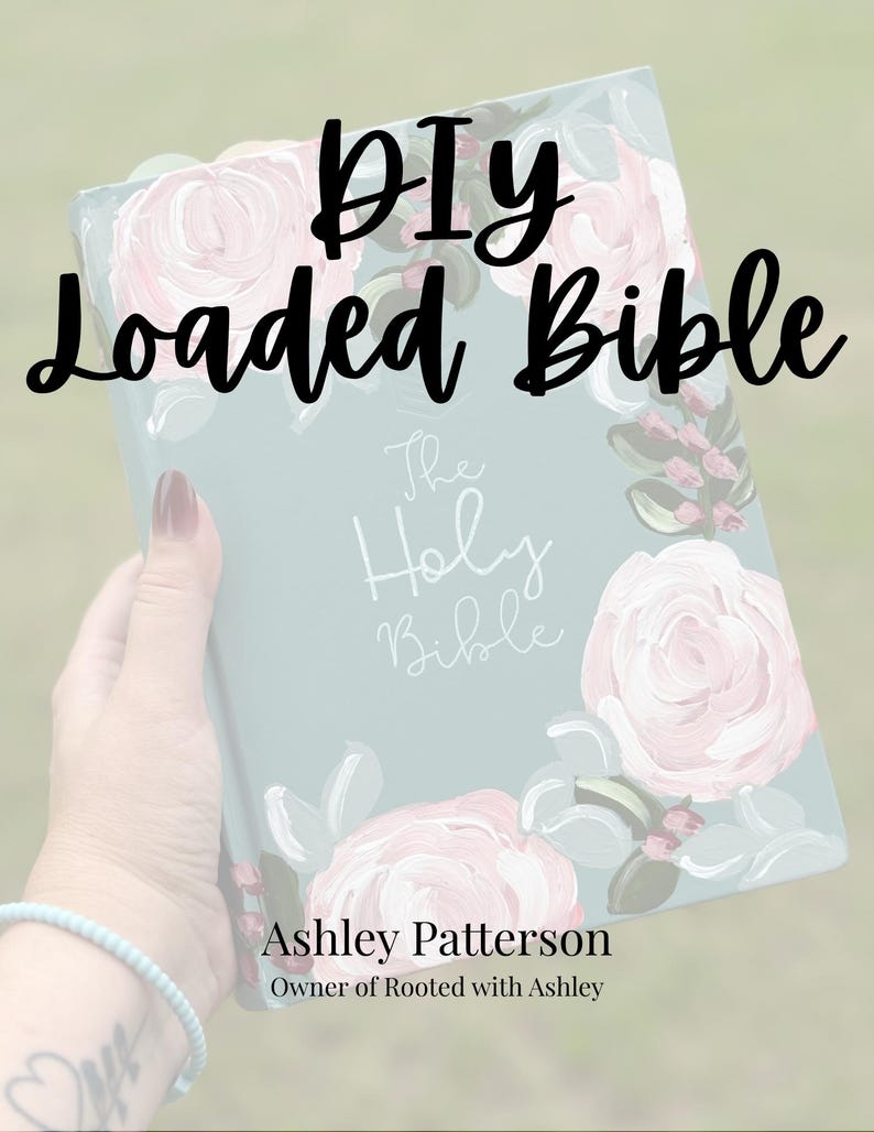 May include: A light blue Bible with a painted floral design featuring pink roses and green leaves. The cover displays the text "The Holy Bible" in white script, with "DIY Loaded Bible" and "Ashley Patterson Owner of Rooted with Ashley" printed.