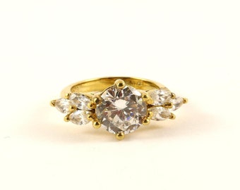 Op de afbeelding: Een gouden ring met een grote, heldere, ronde edelsteen omringd door kleinere, heldere, peervormige edelstenen.