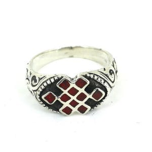 May include: A silver ring featuring an intricate, raised metal band with scrolling patterns and a central oval inlay. The inlay displays a red, geometric knot design set against a dark, recessed background, creating a distinct contrast.