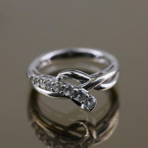 May include: A silver ring with a delicate, intertwined design featuring a row of clear gemstones. The ring has a simple, elegant style.