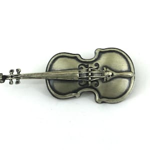 May include: A detailed, antique-style violin brooch in a metallic silver-bronze color. The brooch features intricate detailing of the instrument's body, strings, and scroll, set against a white background. A decorative accessory for music lovers.