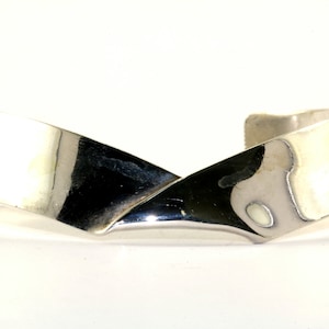 May include: A silver-colored cuff bracelet with a modern, geometric design. The bracelet features a wide, flat band that crosses over itself in the center, creating an X shape. The polished metal reflects light, highlighting its smooth surface.