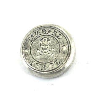 May include: A round, silver-colored metal coin with a skull and crossbones design in the center. The coin has the text "MK BARZ" around the top and "1 TROZ" around the bottom. The coin is on a white background.