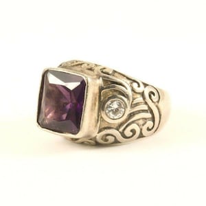 May include: A silver ring with a large, square, purple gemstone. The ring has a decorative, swirling pattern on the band and a small, clear gemstone. The ring is set against a white background.