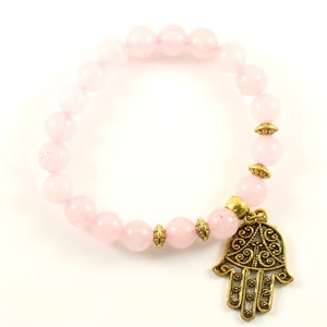 May include: A pink beaded bracelet with a gold-coloured Hamsa hand charm. The bracelet features round, light pink beads and decorative gold-coloured spacers. The Hamsa charm is detailed with an ornate design.