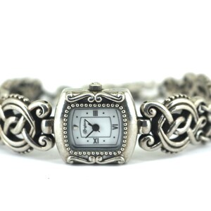 May include: Silver-toned wristwatch with a square face and a white dial. The watch features Roman numerals and a decorative, patterned band. The watch face is approximately 1 inch wide.