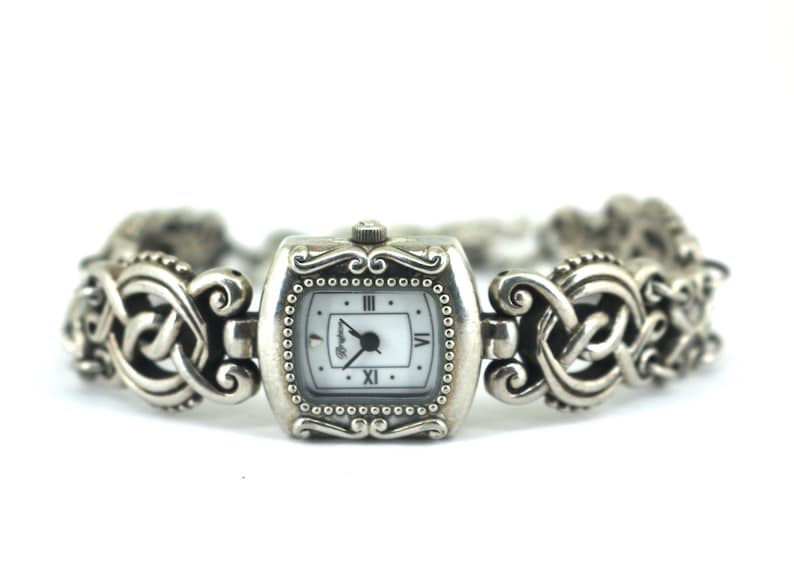 May include: Silver-toned wristwatch with a square face and a white dial. The watch features Roman numerals and a decorative, patterned band. The watch face is approximately 1 inch wide.
