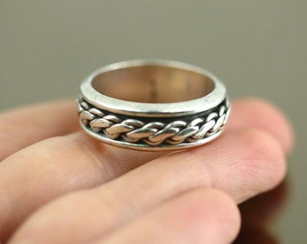 May include: A silver ring with a braided design. The ring is on a person's finger.