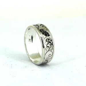 May include: A silver ring with an engraved floral and leaf pattern. The ring has a polished finish and a detailed design that wraps around the band. The black detailing contrasts with the silver metal.