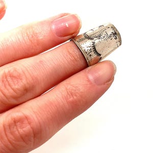 Op de afbeelding: Een zilveren vingerhoed met een decoratief patroon wordt op een vinger gedragen. De vingerhoed heeft een licht aangeslagen uiterlijk, met donkere accenten die het ontwerp benadrukken. Het is een naai-instrument.
